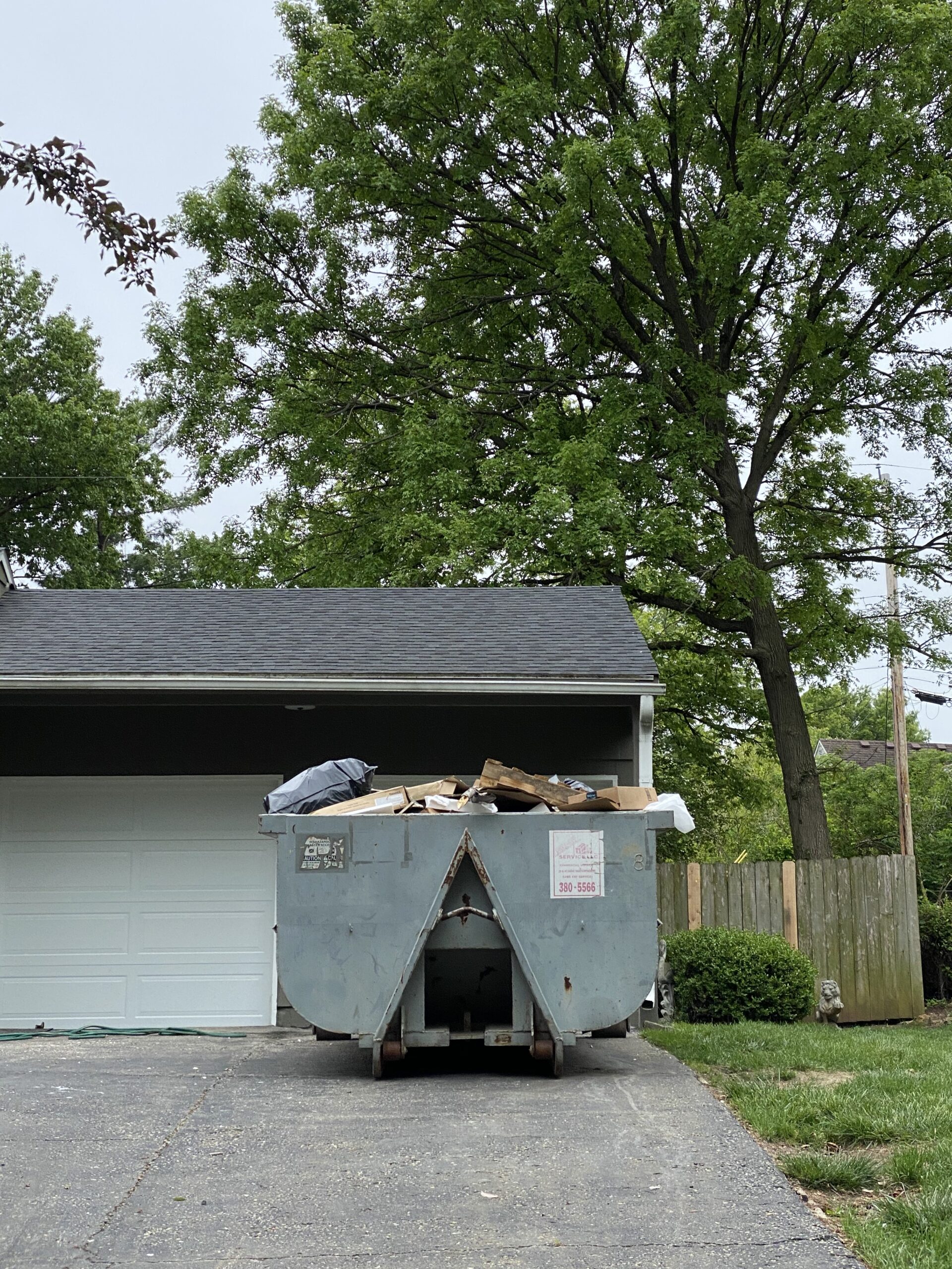Dumpster filled with debris in suburban driveway, surrounded by greenery and residential setting, illustrating junk removal process for decluttering.